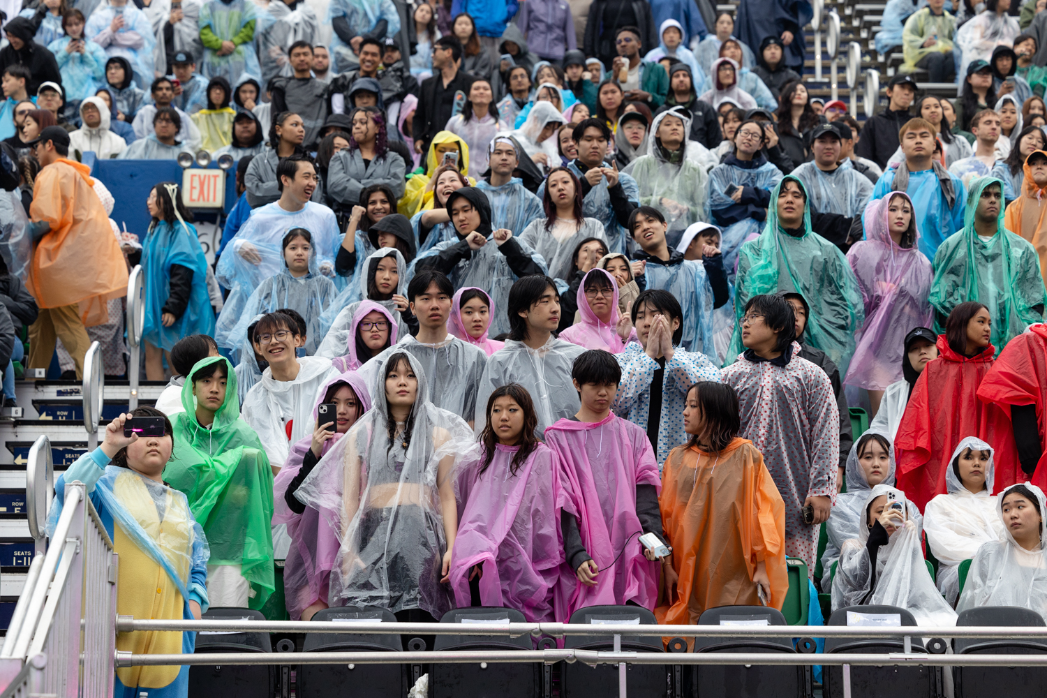 Dozens of people standing and sitting on bleachers — seats which are raised above one another. They are wearing various, colored rain ponchos over their regular clothing.