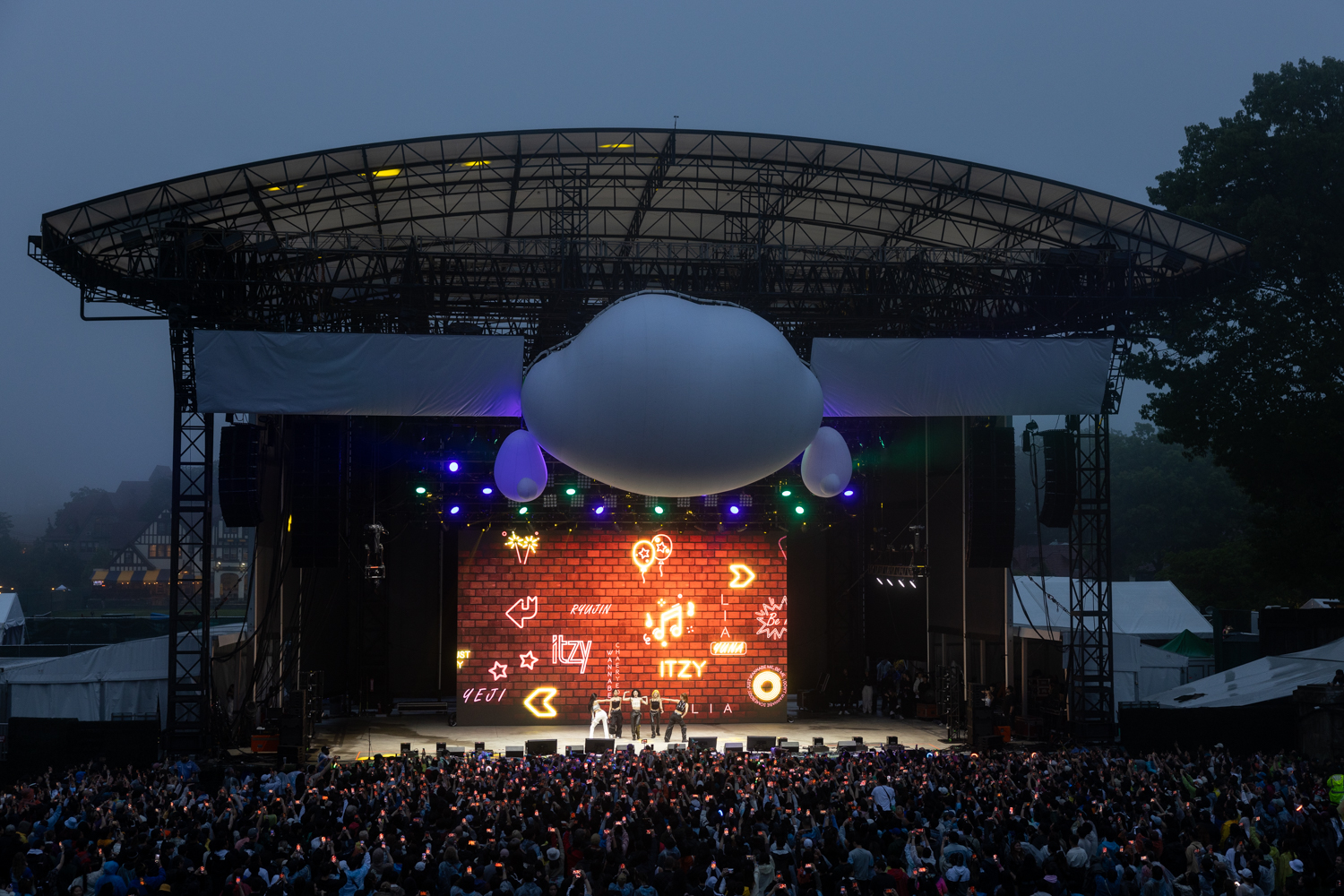 A large crowd of people hold up their phones, recording the five female members of Itzy. Behind them is a screen projecting an image of a brick wall covered in arrows, stars, and various other symbols. Above them on the scaffolding roof of the stage is a large cloud with arms.