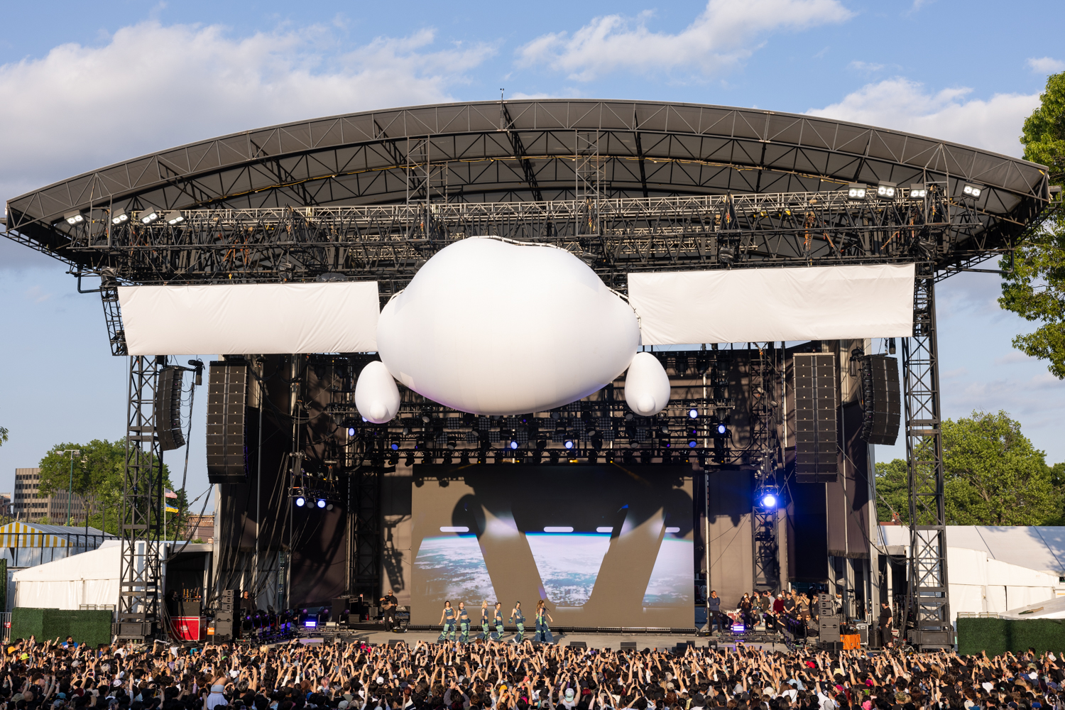 The seven female members of X.G. perform onstage, wearing identical blue and green outfits. They are beneath a large decorative cloud attached to the stage ceiling, in front of a screen displaying a view of Earth from space. A large crowd of people hold up their phones to record the performance.