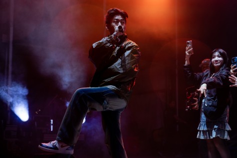 Rich Brian stands on stage with a microphone in his hand under orange and blue hued lights. Behind him, people watch with cellphones in hand.