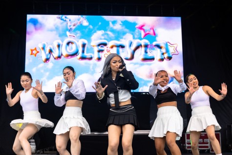 Wolftyla, wearing a black top over a white shirt and a black skirt, holds a microphone on stage as she performs onstage. She is surrounded by two backup dancers wearing white skirts and black-and-white tops. Behind them is a large screen displaying the name “WOLFTYLA” in a bubbly, silver font, surrounded by various colored stars on a sky background.