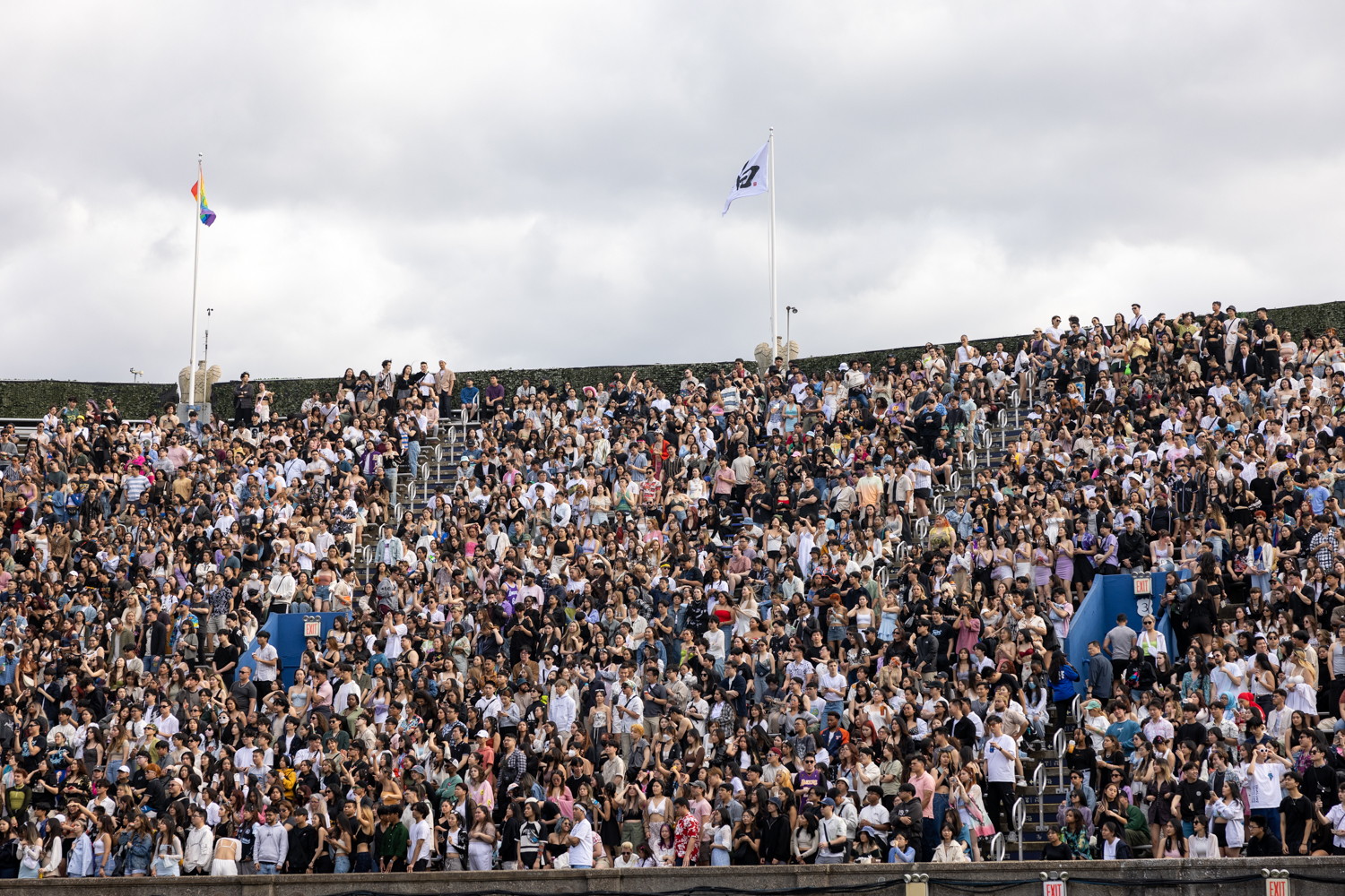 Hundreds of fans sitting and standing in the rising bleacher seats of Forest Hills Stadium. At the top of the seating is green shrubbery and two flag poles.