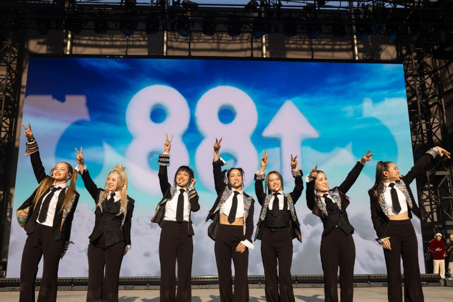 The seven female members of X.G. stand in front of a large screen displaying a white number “88” alongside an upward arrow of the same color over a sky background. They are each wearing identical black suit jackets with rhinestone accents and black ties. The performers make peace signs to the crowd with their hands.