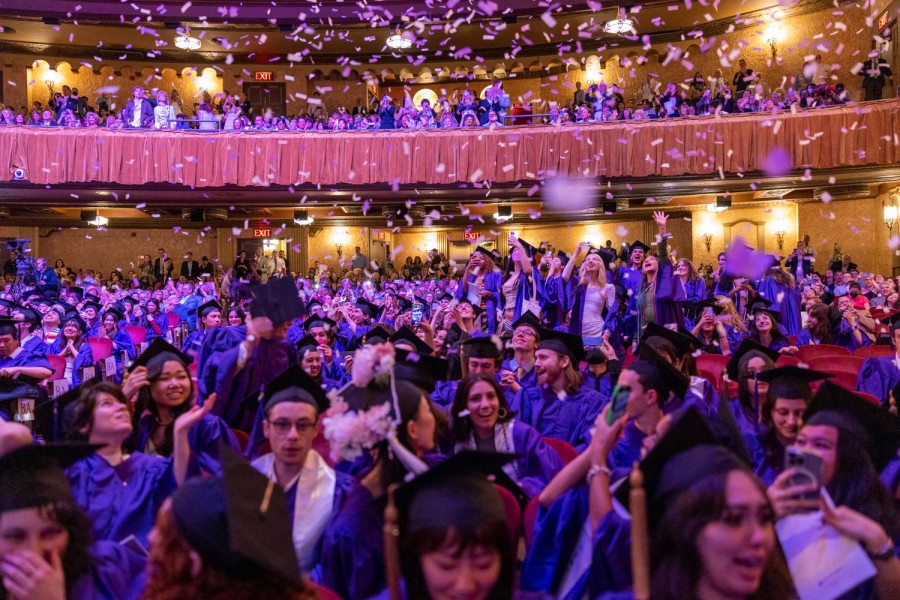 Hundreds of Gallatin students in black caps and purple gowns celebrate their graduation inside of a theater. Purple confetti rains down from above.
