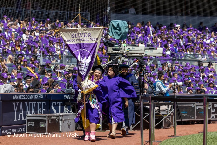 Three people in purple gowns wearing black graduation caps are walking across reddish-brown dirt in a line, with the person in the front smiling with a scarf around her neck made up of photos while simultaneously carrying a large banner which reads SILVER SCHOOL OF SOCIAL WORK and has a logo depicting a half a planet with a cityscape on top of it. Behind the is a large crowd of people sitting in purple graduation gowns and caps, separated from the trio by a wall of netting.