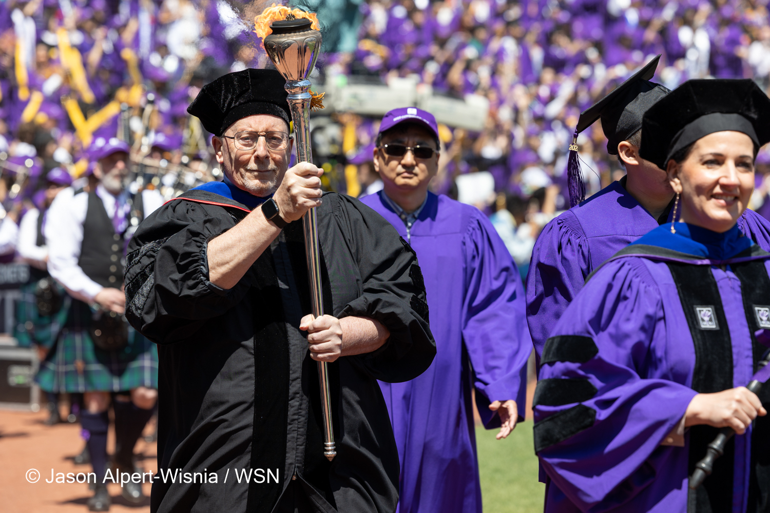 J. Anthony Movshon wearing a black cap and gown walks on the field holding the torch.