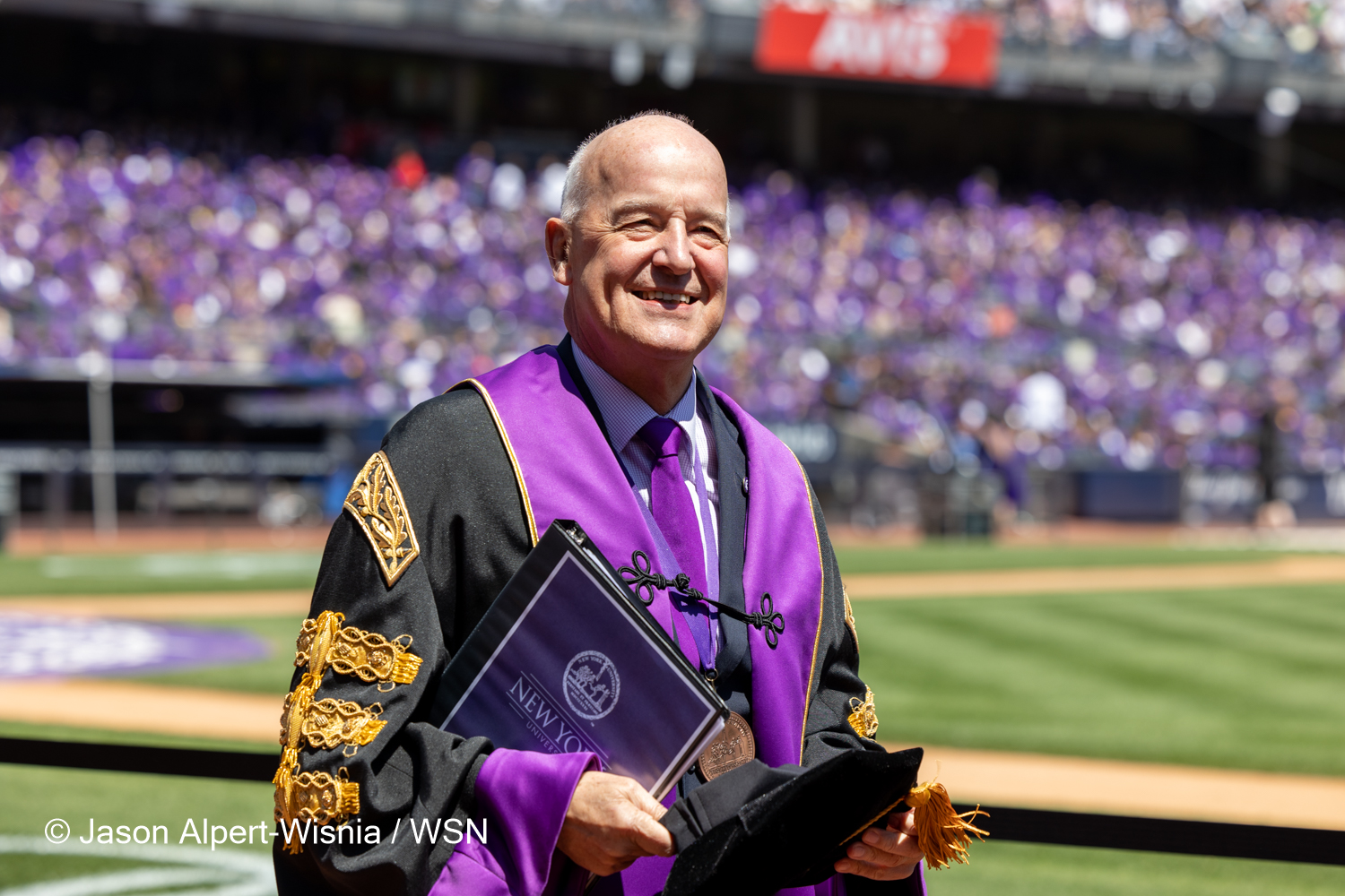 N.Y.U. President Andrew Hamilton walking through a packed Yankee Stadium. He wears a black-and-purple robe with gold embellishments, holding a black graduation cap with a gold tassel and a black binder with purple paper displaying the university’s name and logo.