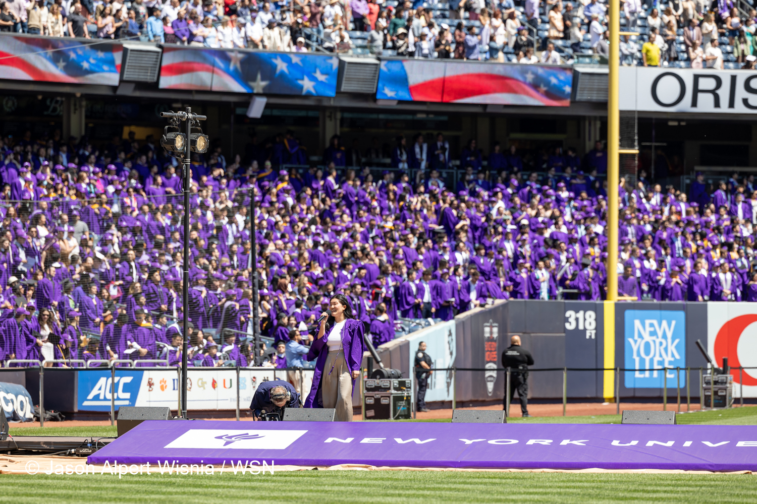 Katie Song-Yi Nam wearing a purple gown and white shirt sings the national anthem on the field.