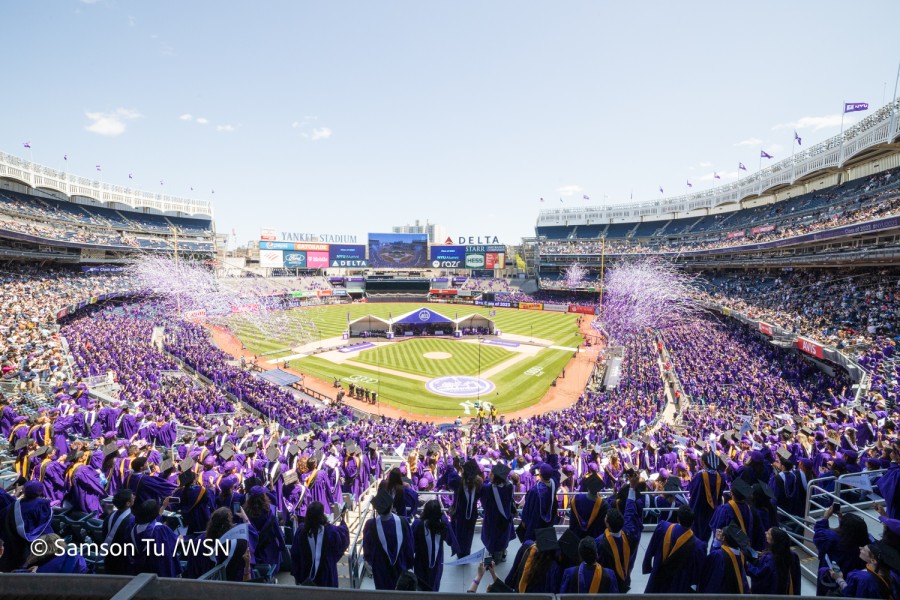 A wide-angle view of Yankee Stadium whose stands are packed with N.Y.U. graduates dressed in purple gowns. Purple and white confetti flies in the air.