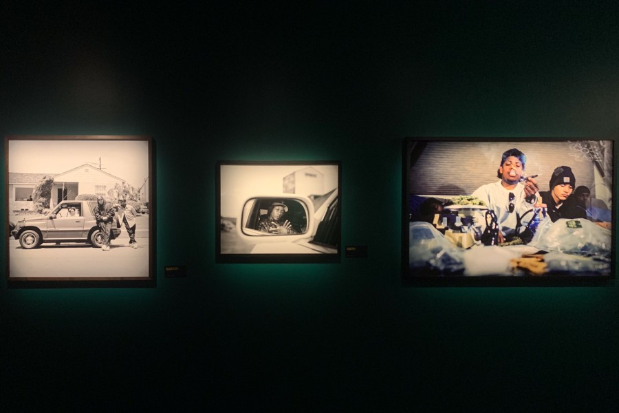 Three photographic prints hang on a green wall. To the left is a black-and-white photo with a car and two people standing next to it; in the center is a black-and-white photo of a person looking into a rearview mirror; on the right is a photo of a person smoking a cigarette with people sitting next to him.