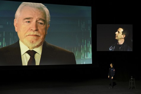 In a still from “Succession,” in an auditorium, an image of Logan Roy is projected on a screen behind Kendall Roy, who is looking up at the screen.