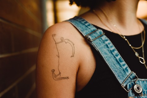 A shoulder tattoo done by Ella Bowie on C.A.S. first-year Ava Santiago. The tattoo shows two people, one sitting and one standing, bending over to meet the other’s level, holding a tall flower together.