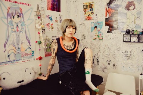 J. sits on a tattoo bench in their studio. They are facing the camera, and behind them, the walls of their studio are covered in posters and their work.