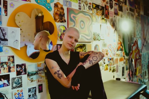 Phoebe Satterwhite sits on a tattoo bench in their personal studio. They are facing the camera. The walls behind them are covered in trippy, multicolored art pieces, some of which were made by Satterwhite.