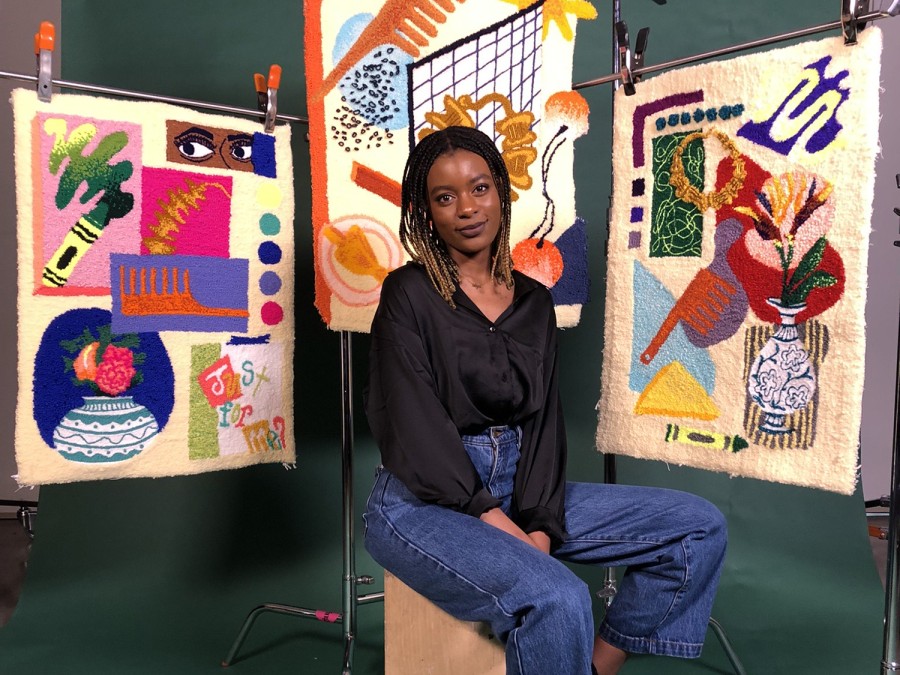 Aliyah Salmon, wearing a black shirt and jeans, sits on a wooden box in front of a dark green backdrop. Three pieces of tapestry art hang from metal scaffolding behind her. They depict images of objects like a green crayon, a blue-and-white vase and eyes.