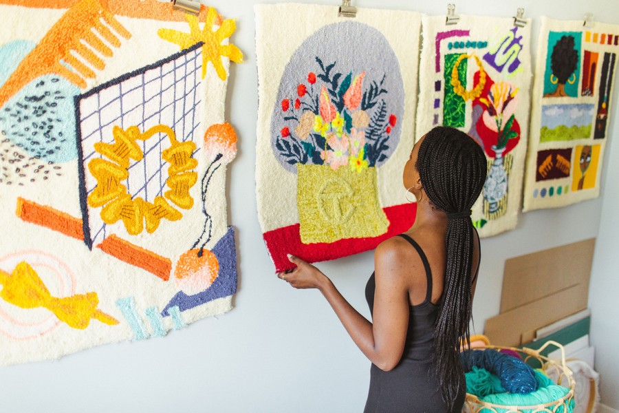 Aliyah Salmon, wearing a black shirt, hangs a piece of rug art on her wall depicting a yellow Telfar bag with various colored flowers coming out of it. It is besides various other pieces of art of the same medium.