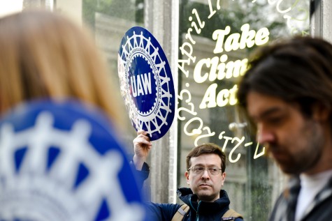 A man holds a blue, circular “U.A.W.” sign. He is looking to his side.