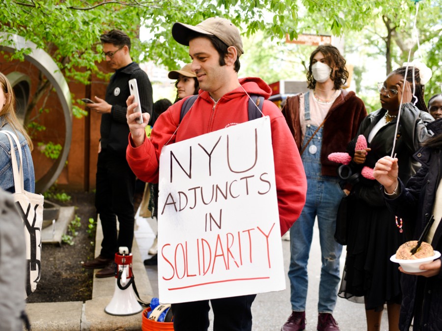 A person wearing a red sweatshirt holding a cardboard sign that reads “N.Y.U. Adjuncts in Solidarity.” He is at a rally next to the Bobst Library and is holding his phone, taking a picture. There is a crowd of people around him.