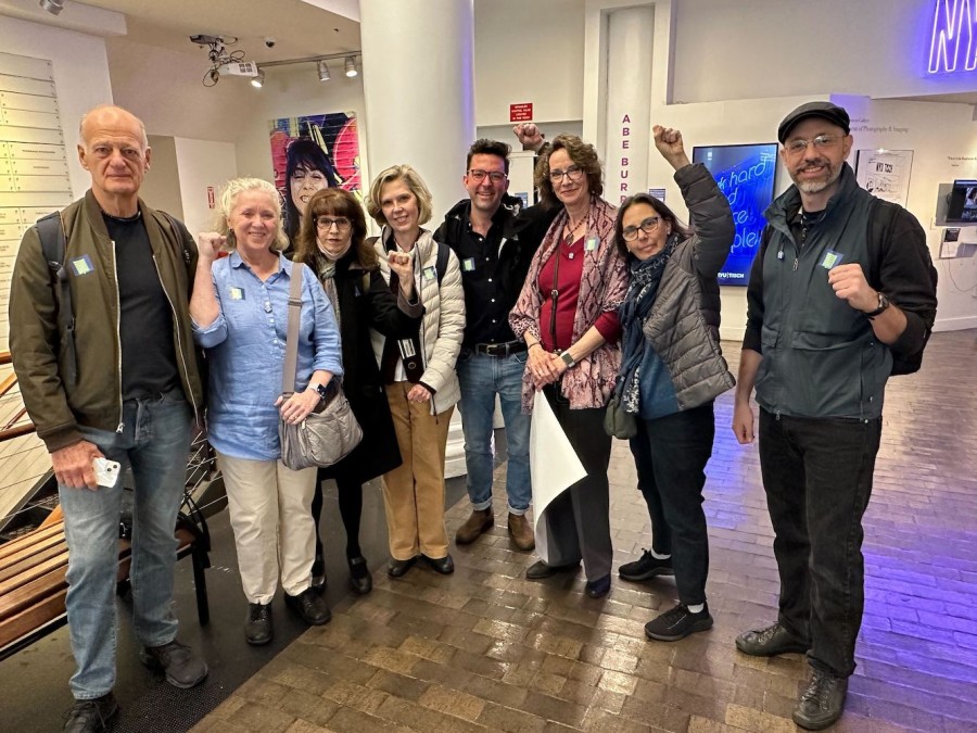 Professors stand in the lobby of a Tisch School of the Arts building, smiling and holding up their fists. From left to right: Jack Eppler, Melanie Vaughan, Michelle Rosen, Jill Edwards, Robert Frost, Carolyn Paulus, Elizabeth Olesker and Gerasimos Karavitis.