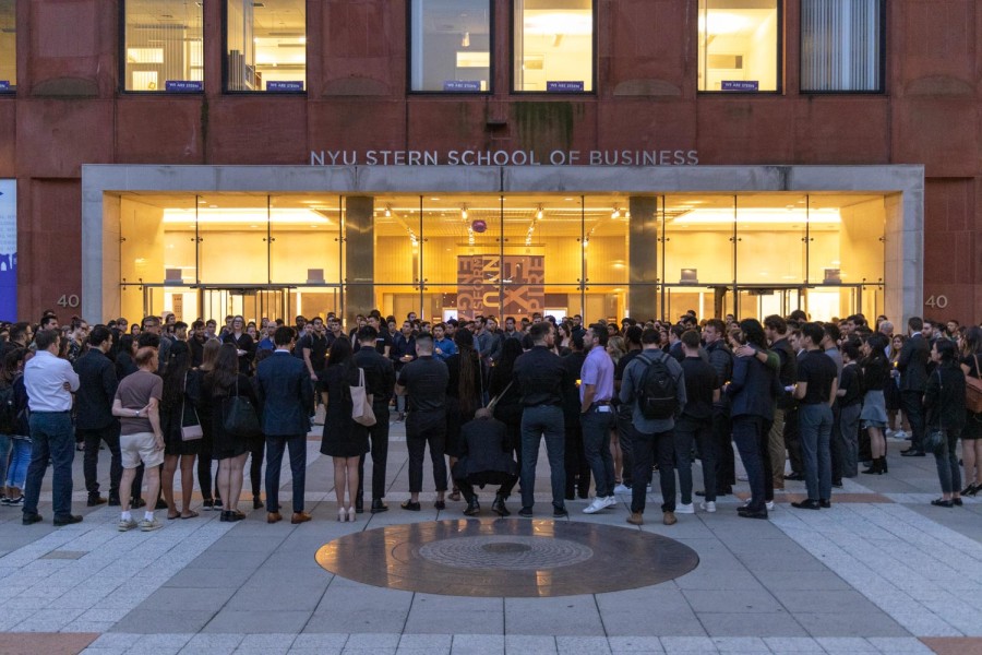 More than one hundred people dressed in black gather in a circle during a candlelight vigil in a plaza in front of a red sandstone building.