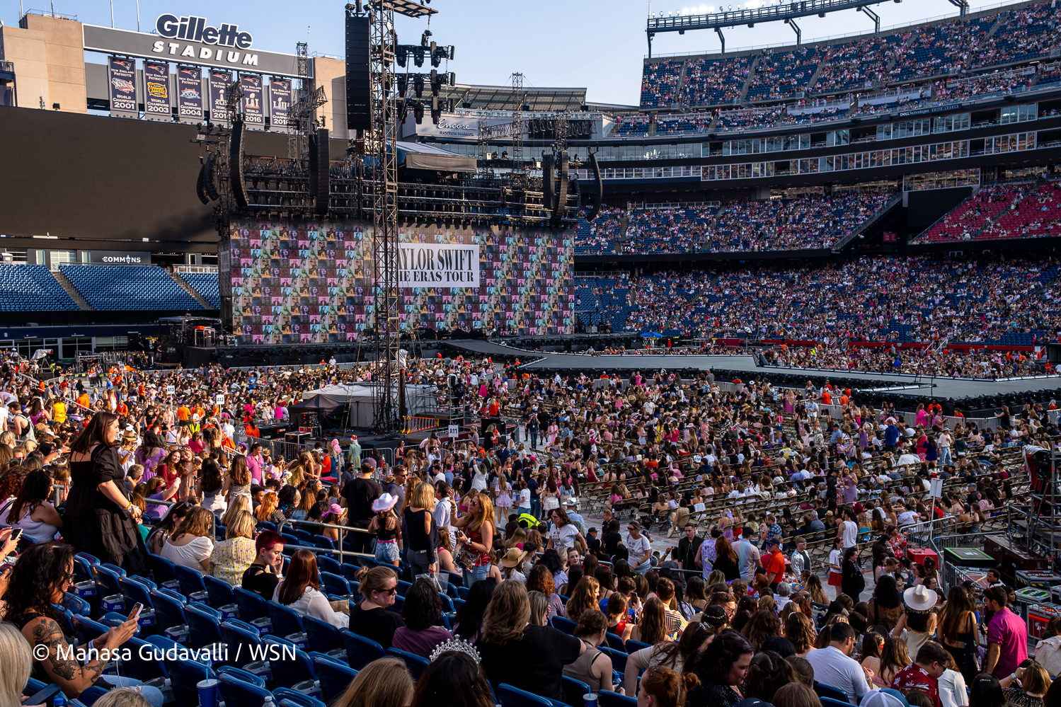 A large crowd fills the floor and stands of Gillette Stadium, surrounding a stage and a tower at the center of the stadium. The stage has a background featuring colorful photos of Taylor Swift, with the text “TAYLOR SWIFT THE ERAS TOUR” overlaid in black on a white rectangle. A sign reading “Gillette Stadium” overlooks the crowd from the top of the stadium.