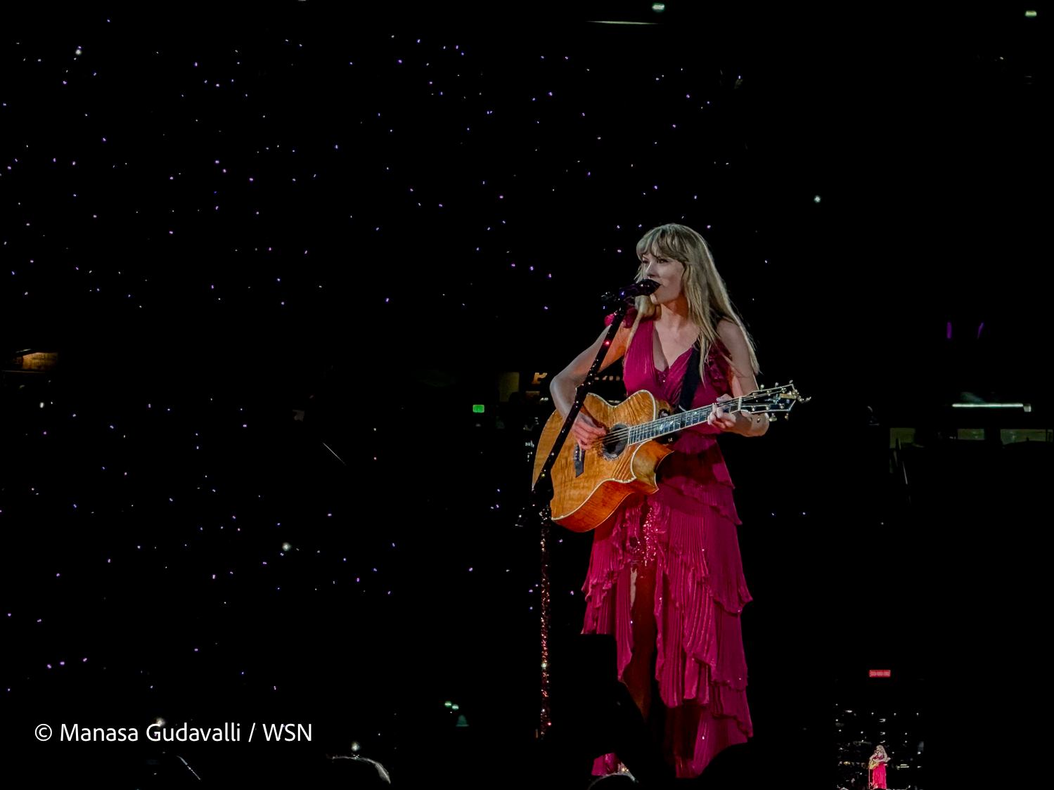 Taylor Swift performs before a screen background that is black with white stars, standing with a guitar in hand. Swift wears a hot pink mid-length dress with layered ruffles as she sings into a standing microphone.
