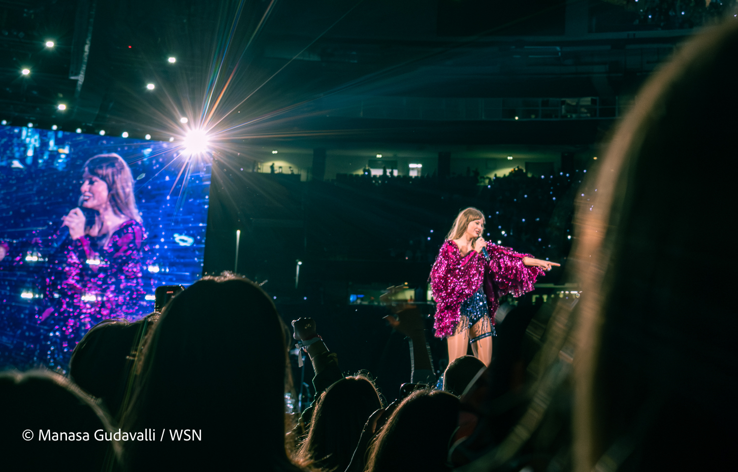 Taylor Swift points out into the crowd at Gillette Stadium with a microphone in hand, wearing a sparkly black leotard with a sparkly hot pink top that has long ruffled sleeves. In the background, fans hold up their phones in the mostly dark stadium, with fans facing the stage standing in the foreground. A screen playing live footage of Swift’s performance stands behind her along with a bright white light.