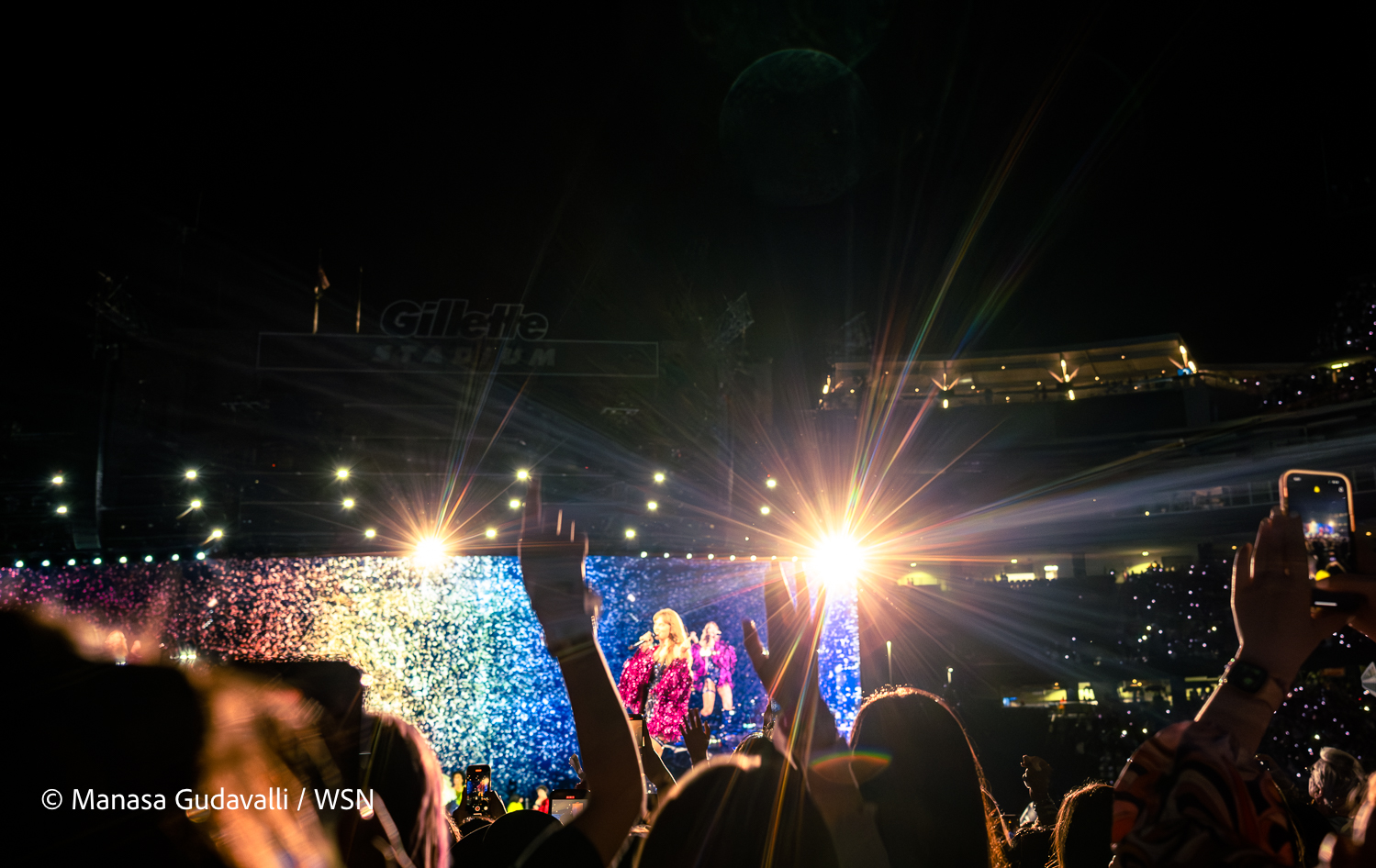 Taylor Swift performs before a crowd on the ground level of Gillette Stadium, wearing a sparkly pink and black leotard with long pink sleeves. Fans raise their hands up in the air as Swift performs in front of a background displaying live footage of her performance. Two bright yellow-white lights shine behind her.