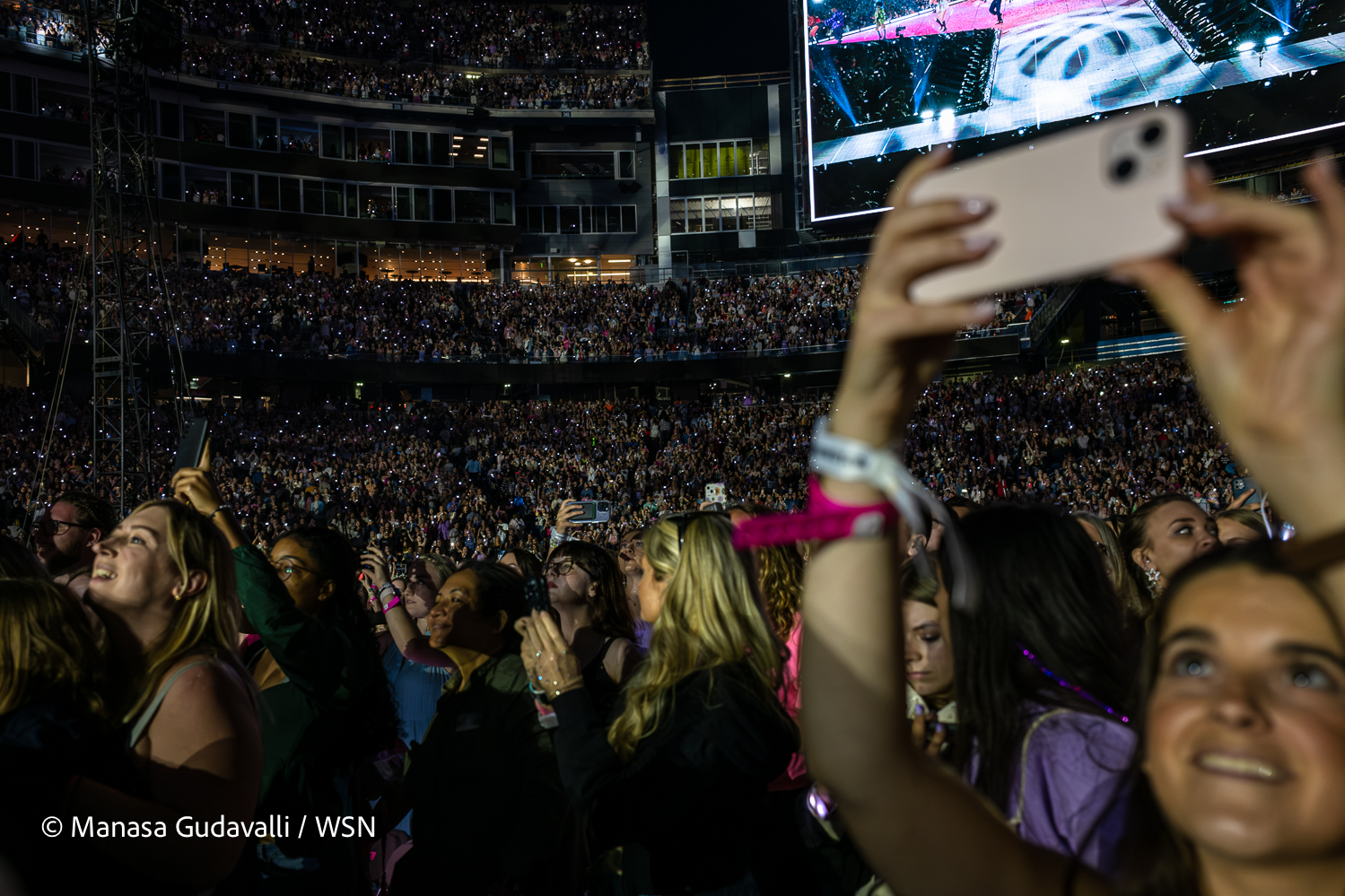 The crowd watches Taylor Swift’s performance. A woman raises her phone to film the performance. Some concert attendees shine the flashlight on their phones toward the stage.