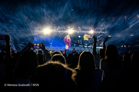 Taylor Swift, wearing a purple feather jacket, a pair of black shorts and boots, and a backup dancer, wearing a shiny golden jacket, sings on a stage filled with rainbow-colored confetti. Concert attendees clap along and film the performance.