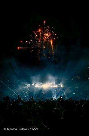 Taylor Swift waves toward the audience against a backlit stage. Above in the night sky are orange fireworks.