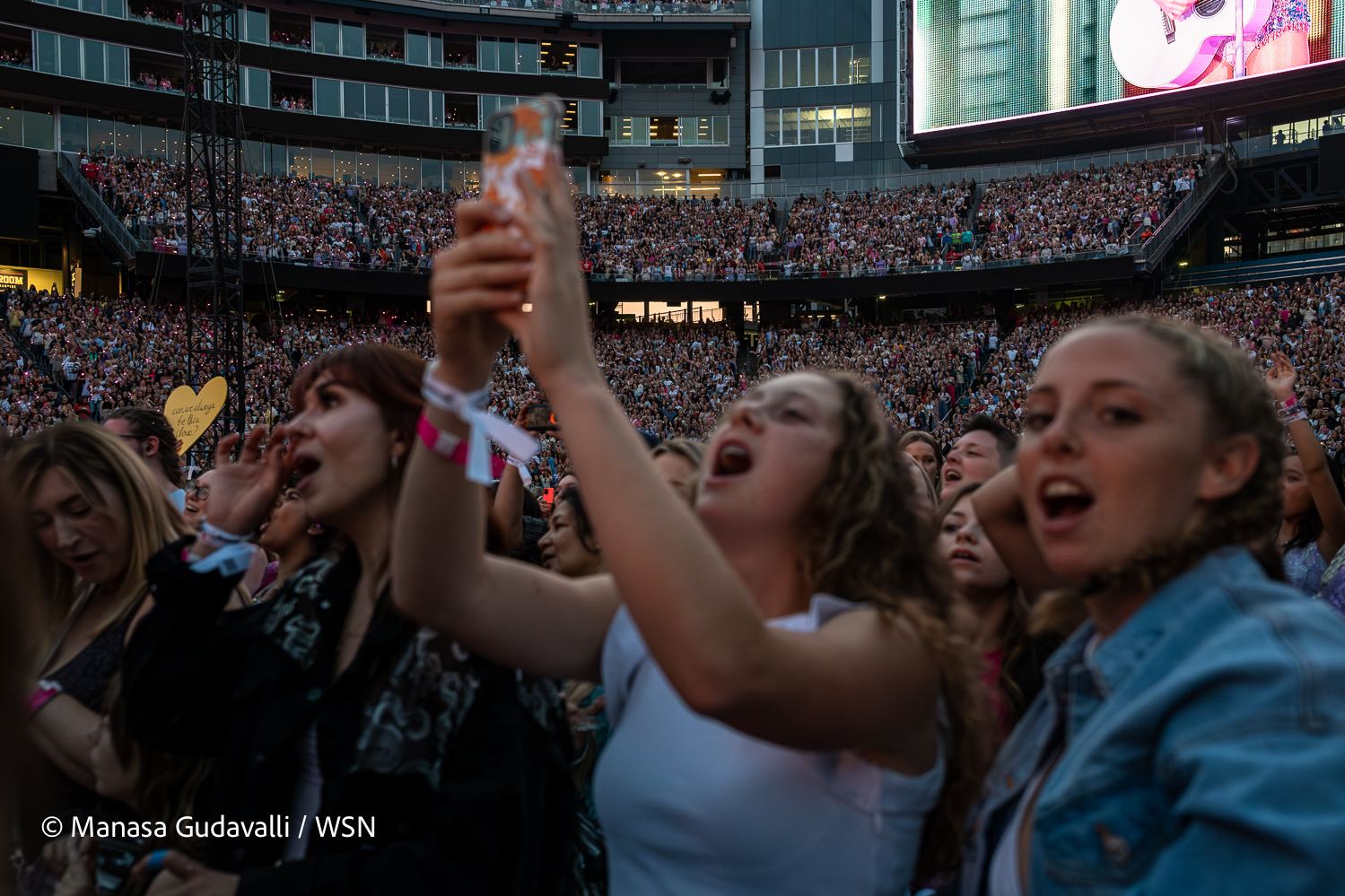 The crowd sings along to Taylor Swift’s performance. A woman in white raises her phone to film the performance.