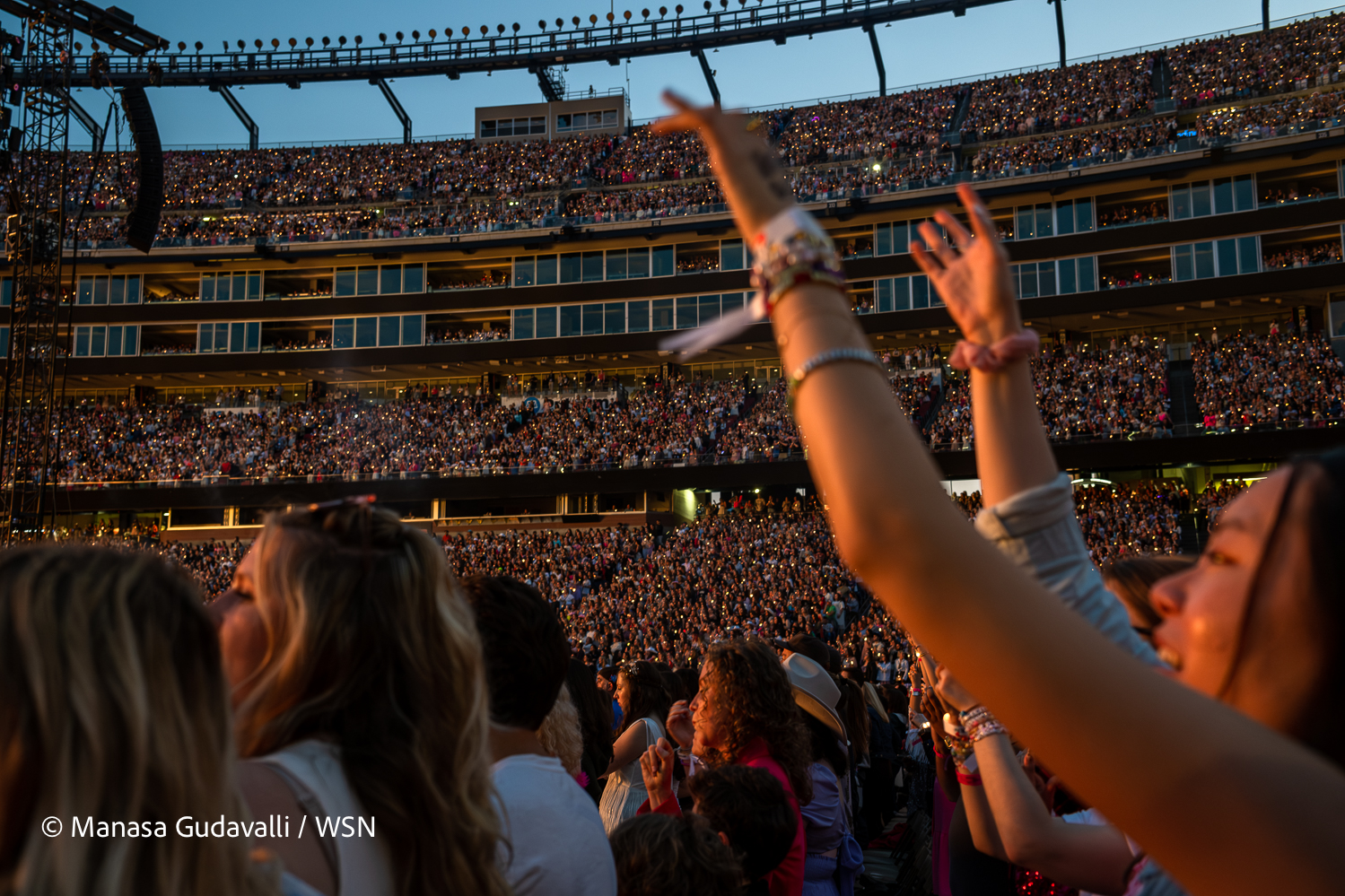 Fans fill the stands of Gillette Stadium as Taylor Swift performs, a yellow-white light shining on them from the stage. In the foreground, a fan wearing multiple bracelets raises their arms toward the stage.
