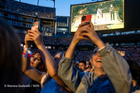 Concert attendees film Taylor Swift’s performance with their phone. In the background is a jumbotron showing Taylor Swift in a white dress with a guitar strapped to her shoulder.