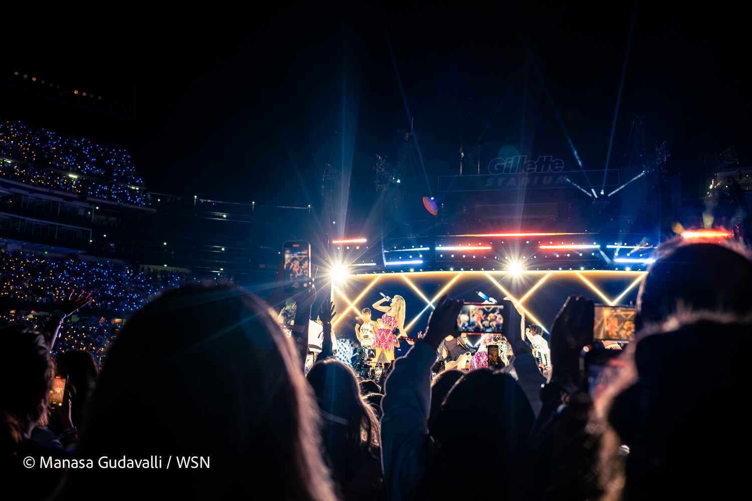 Taylor Swift, wearing a pink shiny top and skirt, sings into a microphone as concert attendees film her performance.