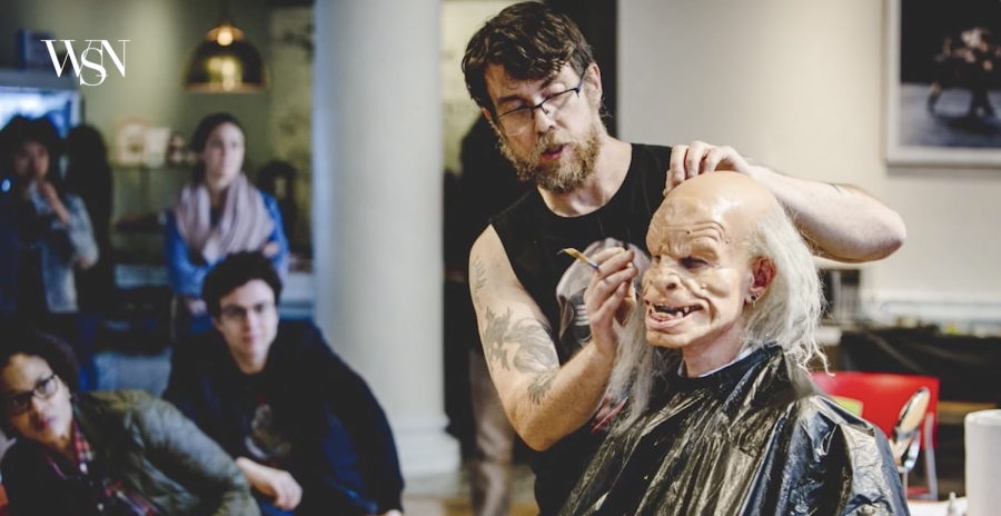 Rob Benevides applies a white wig to an actor wearing a prosthetic costume face.