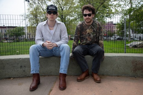 Will O’Connor and Matt Connelly, both wearing sunglasses, sit on a stoop in front of wire fencing. Behind them in the distance are the benches and trees of a public park.