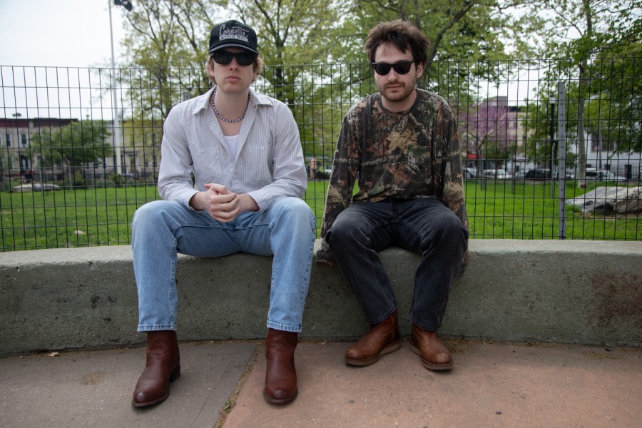 Will O’Connor and Matt Connelly, both wearing sunglasses, sit on a stoop in front of wire fencing. Behind them in the distance are the benches and trees of a public park.