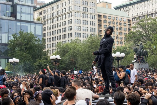 A young man wearing all black and holding a cane stands on top of a car. A large crowd of hundreds of young people stands behind him in Union Square.