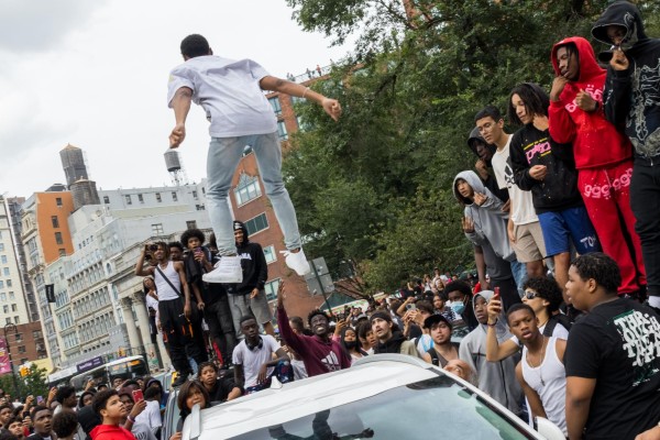 A young man wearing light clothing jumps about two feet in the air above the roof of a white car. The crowd behind him looks on; many are visibly surprised.