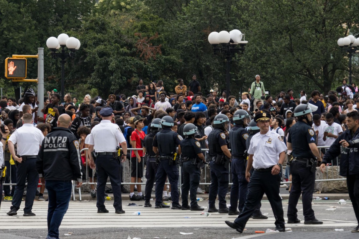 A line of police officers in riot helmets stand in a crosswalk. Behind them, metal barricades contain a dense crowd of young people.