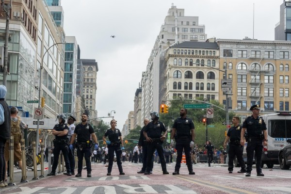 A line of police officers stand side by side in a city street. A quadcopter drone hovers above them.