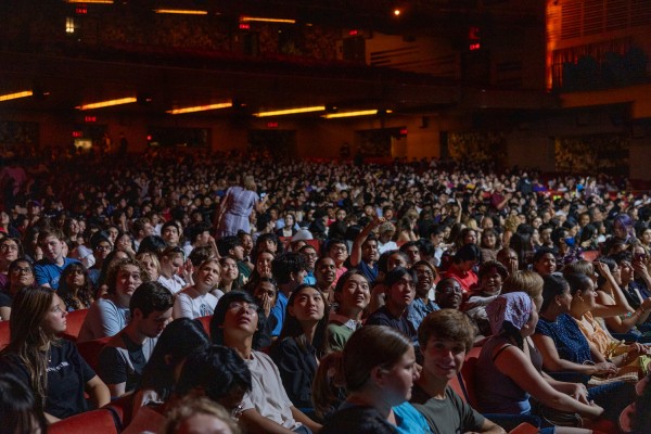 An audience of hundreds are seated in the orchestra level of a large theater, looking up toward a white light. Yellow lights are visible in the background.