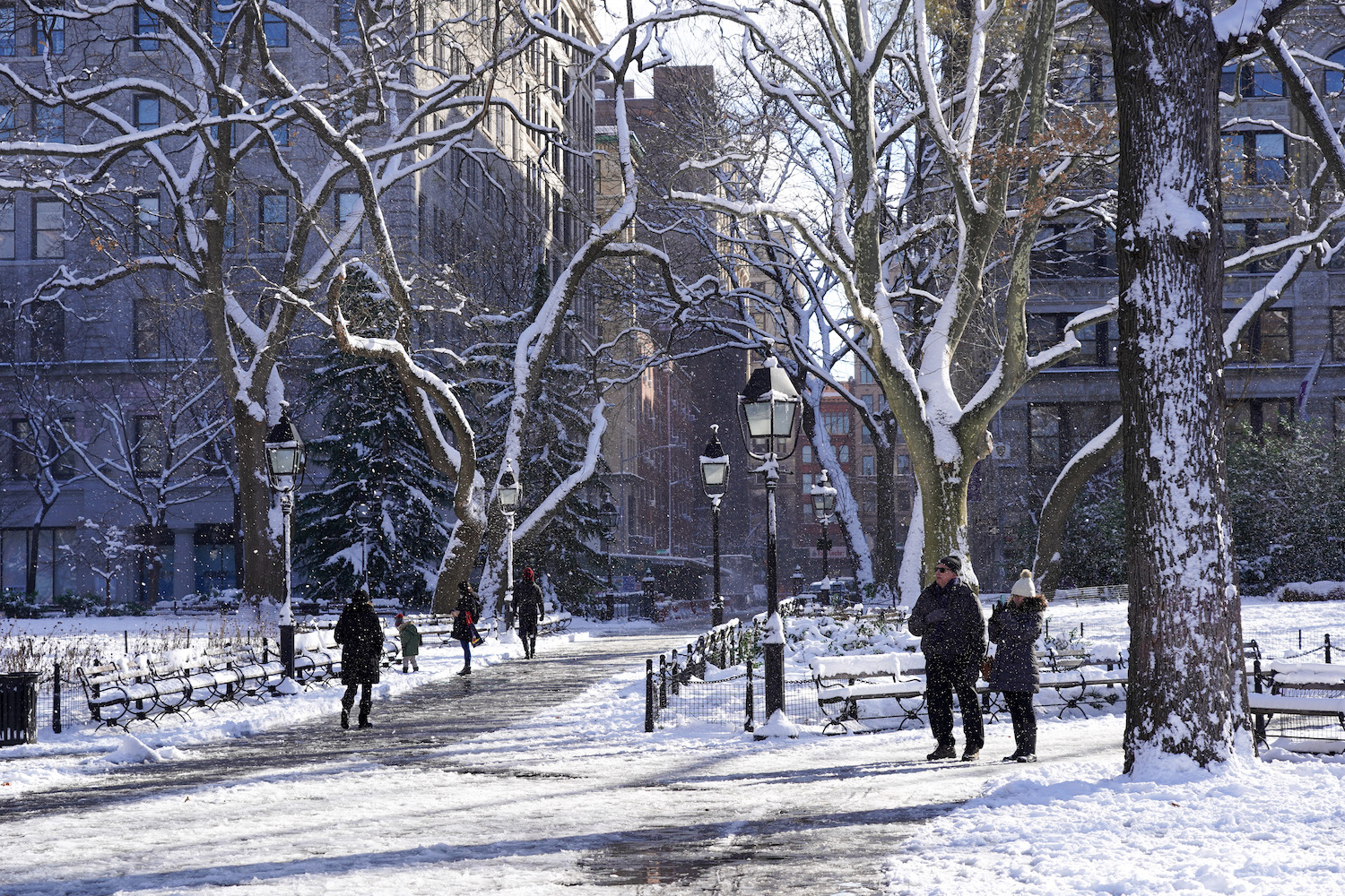 Pedestrians dressed in heavy jackets walk on the east side of Washington Square Park covered in snow.