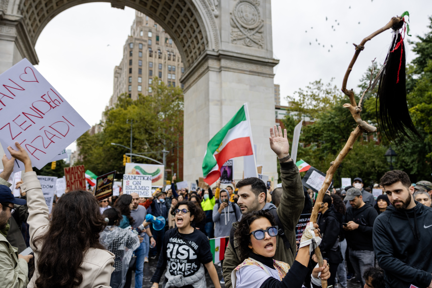 A crowd of protesters standing under Washington Square Arch holding the Iranian flag and signs with a person holding a wood stick with a strand of black hair hanging by the end of it.