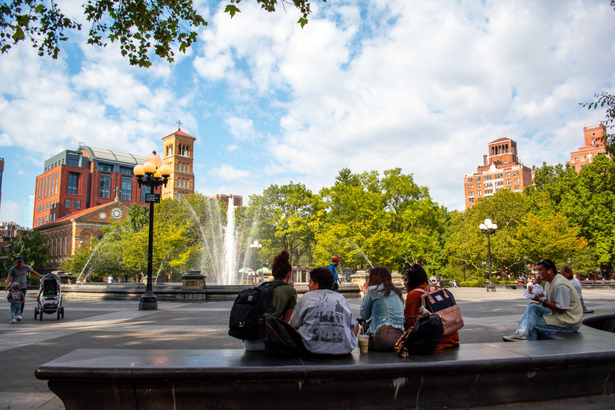 A wide-angle view of the fountain in Washington Square Park. Parkgoers sit on the bench in the foreground.