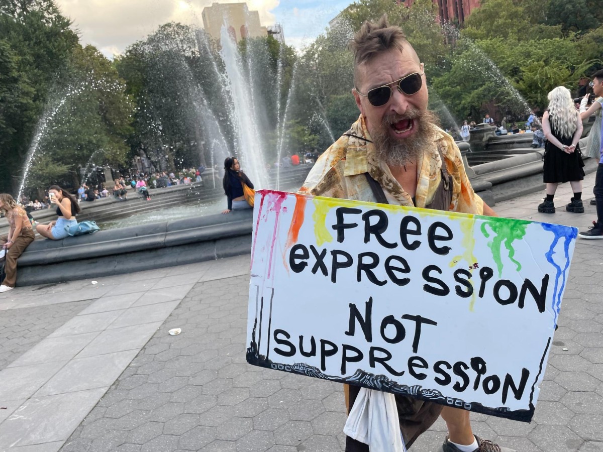 A man stands in front of the fountain at Washington Square Park holding a sign labeled “Free Expression Not Suppression” with his other hand outstretched in a fist. A police officer dressed in uniform stands to the man’s right, facing him.