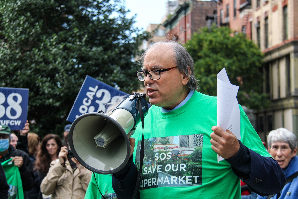 Alan Gerson wearing a green t-shirt with texts that read “S.O.S. Save our Supermarket” speaks to a crowd with a megaphone.