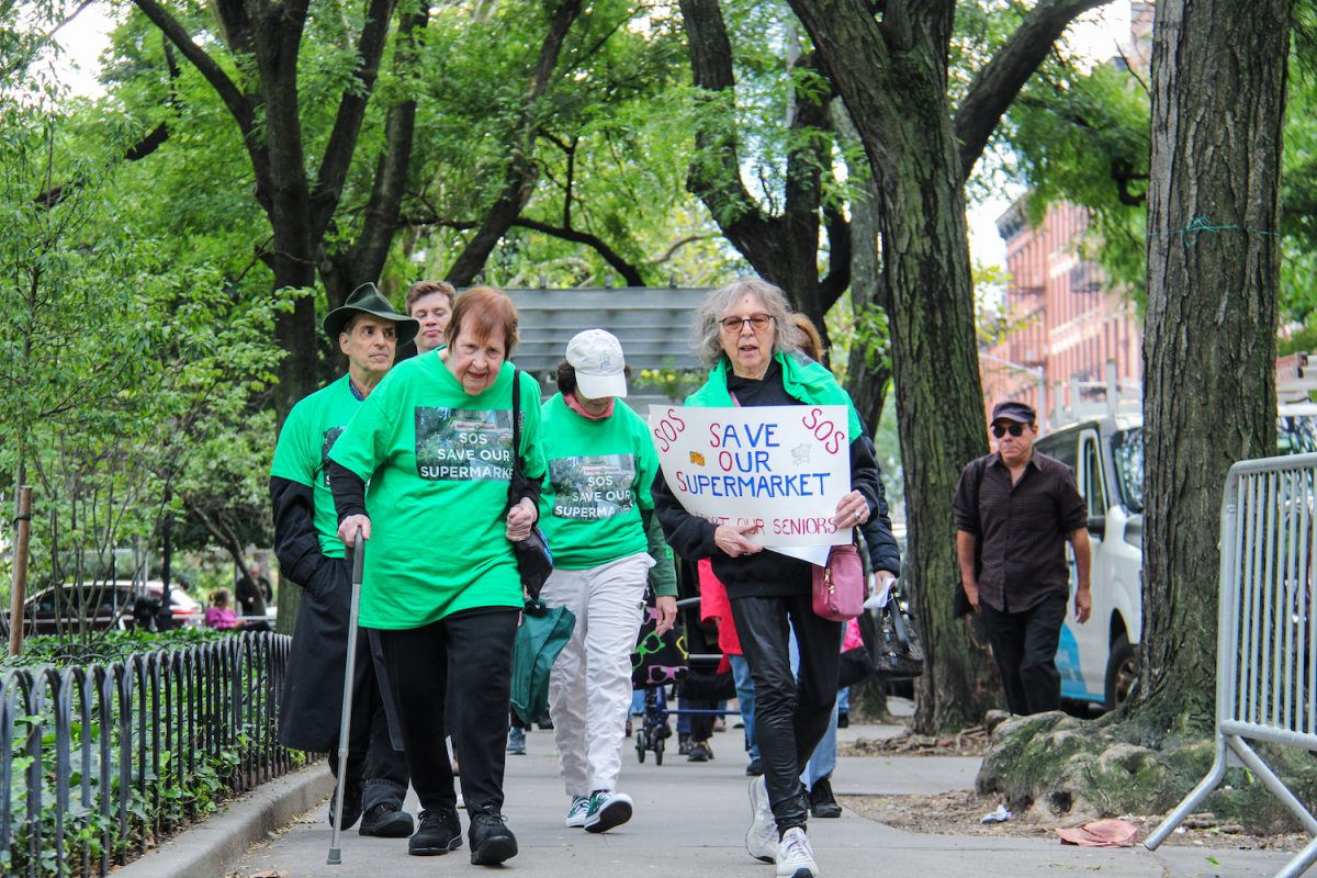 A group of protestors wearing green shirts with the words “S.O.S. SAVE OUR SUPERMARKET ” printed on them, march along a sidewalk. A woman on the right holds a sign that says “SAVE OUR SUPERMARKET”.