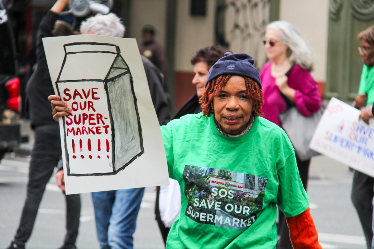 A woman wearing a green shirt and a navy blue hat holding a sign that says “SAVE OUR SUPERMARKET.”